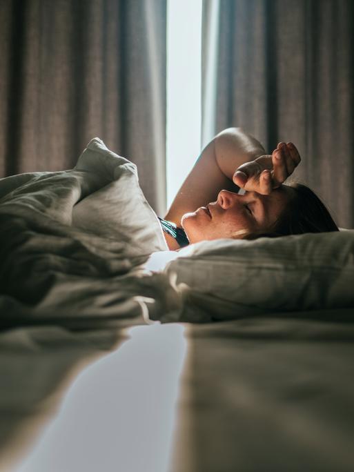 Eine Frau liegt im Bett. Sonniges Tageslicht fällt durch die Vorhänge im Hintergrund. Sie hatte eine Hand an die Stirn gelegt. Eine Frau liegt im Bett. Sonniges Tageslicht fällt durch die Vorhänge im Hintergrund. Sie hatte eine Hand an die Stirn gelegt.