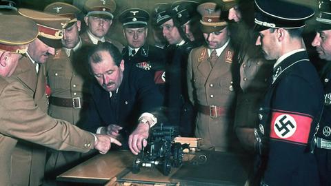 Ferdinand Porsche (1875-1951) zeigt auf ein Automodell, mehrere Männer stehen in Uniformen um ihn herum, darunter Hitler