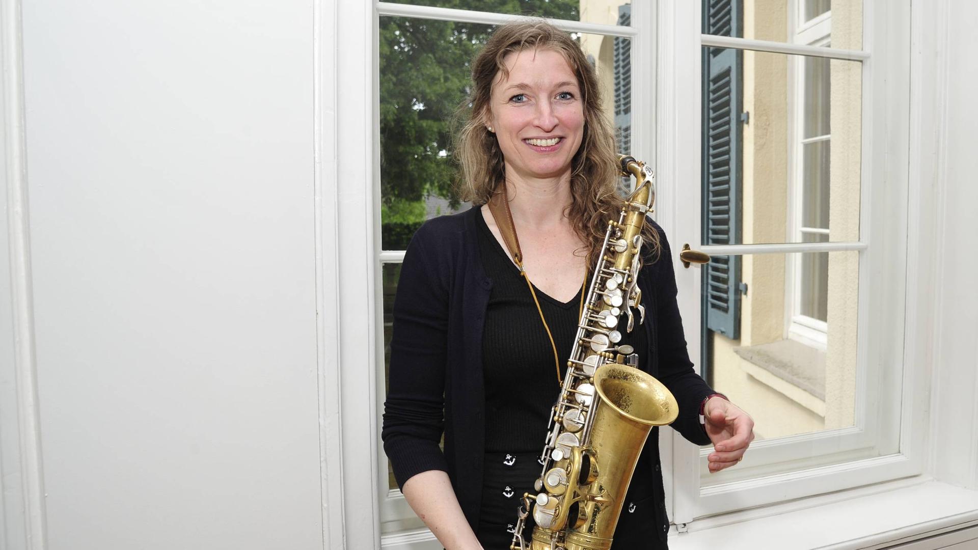 Eine Frau, Nicole Johänntgen, steht vor einem Fenster einer Villa. Sie trägt schwarze Kleidung und hat ein Saxofon in der Hand. 