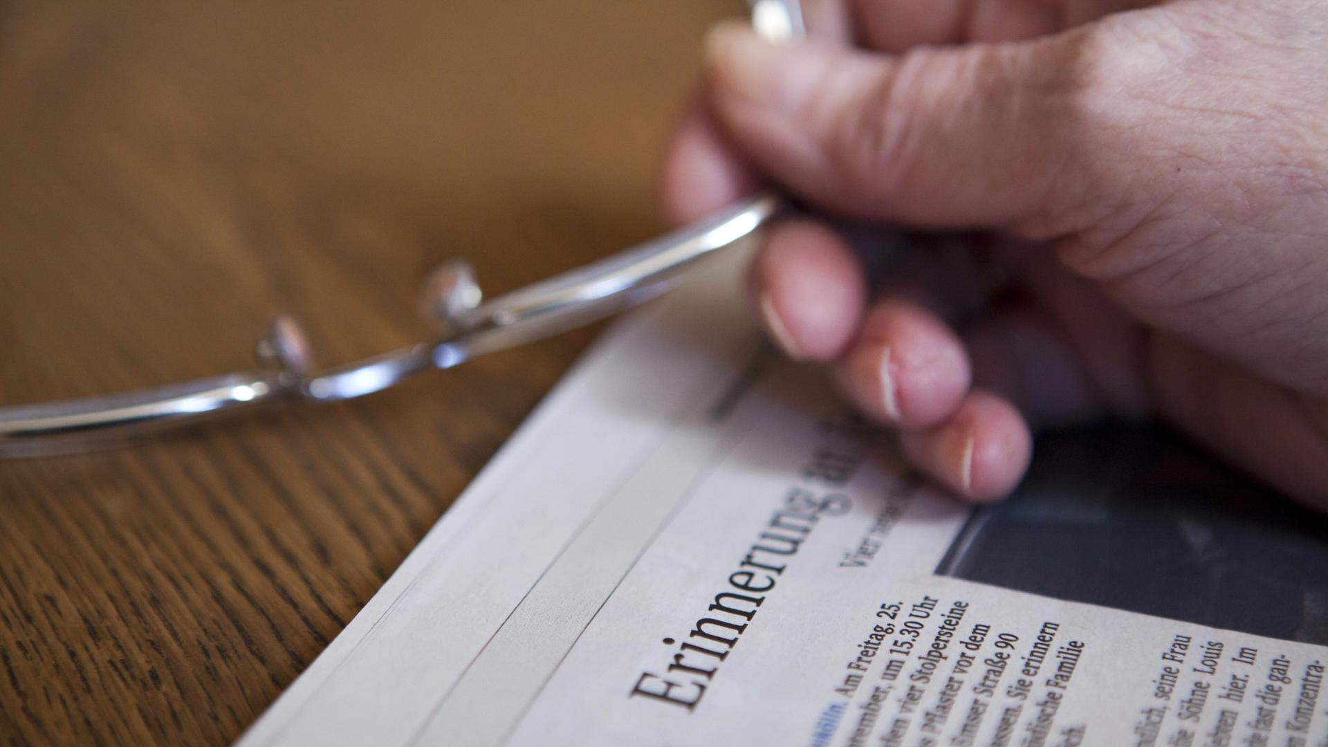 Die Hand einer Seniorin hält eine Brille in der Hand über einer Zeitung, in der im Titel das Wort "Erinnerung" zu lesen ist (gestellte Szene). Die Hand einer Seniorin hält eine Brille in der Hand über einer Zeitung, in der im Titel das Wort "Erinnerung" zu lesen ist (gestellte Szene).