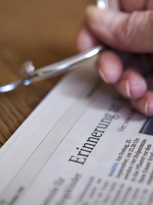 Die Hand einer Seniorin hält eine Brille in der Hand über einer Zeitung, in der im Titel das Wort "Erinnerung" zu lesen ist (gestellte Szene).