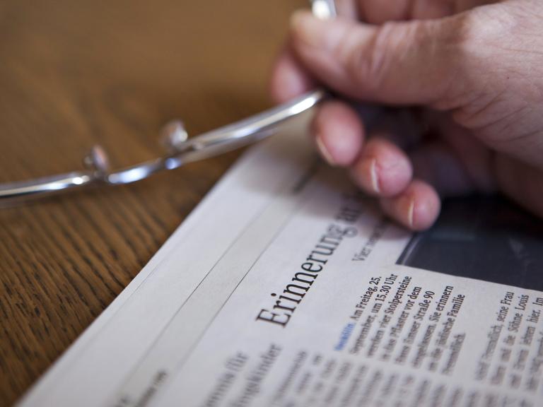 Die Hand einer Seniorin hält eine Brille in der Hand über einer Zeitung, in der im Titel das Wort "Erinnerung" zu lesen ist (gestellte Szene).