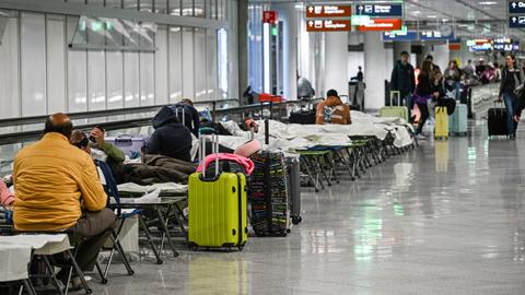 Das Bild zeigt Menschen, die in den Gängen eines Flughafens auf Feldbetten sitzen oder unter Decken liegend schlafen. 
