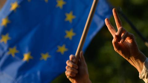 Eine Hand macht ein Peace-Zeichen, im Hintergrund weht eine Europaflagge