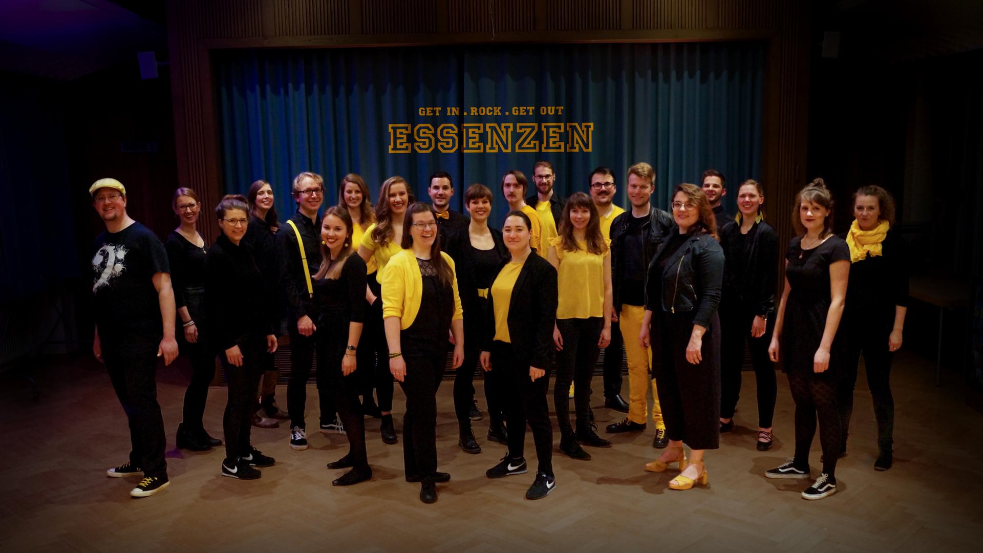 Gruppenbild des Chores vor einem dunklen Vorhang. Die Sängerinnen und Sänger tragen dunkle Chorkleidung und zum Teil gelbe Oberteile.