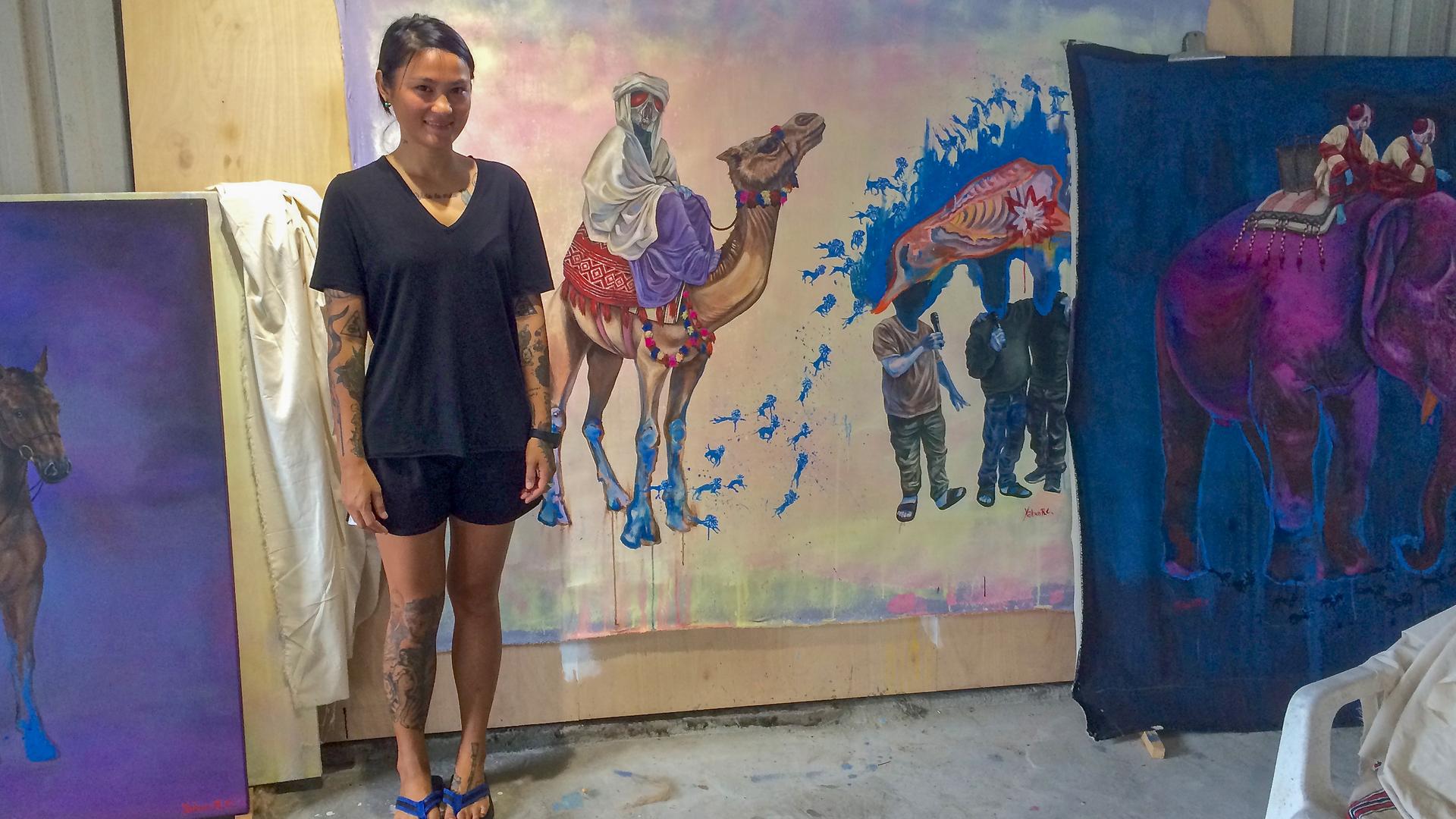 Eine junge Frau steht vor großformatigen Gemälden bunter Kamele.