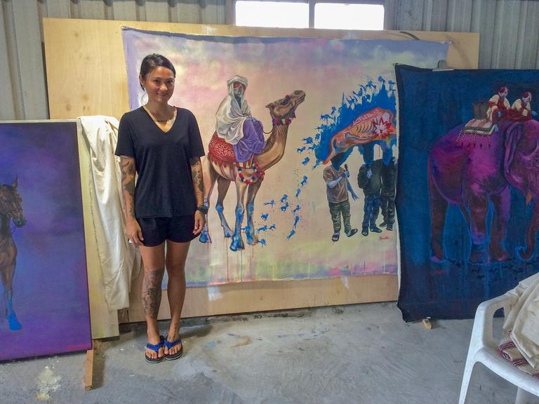 Eine junge Frau steht vor großformatigen Gemälden bunter Kamele.