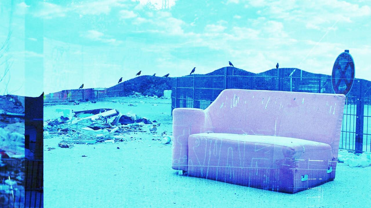 Ein rosa Sofa auf einer Art Müllkippe, im Hintergrund Berge, blau eingefärbt
