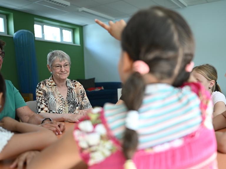 Zwei Seniorinnen sitzen mit Kindern an einem Tisch in einer Grundschule.