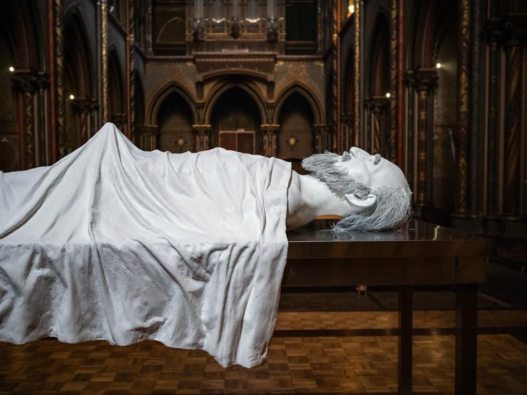 Eine Kunstharz-Figur liegt auf einem Metallbett unter einem weißen Laken. In der Mitte des Körpers steht das Laken nach oben ab.
      