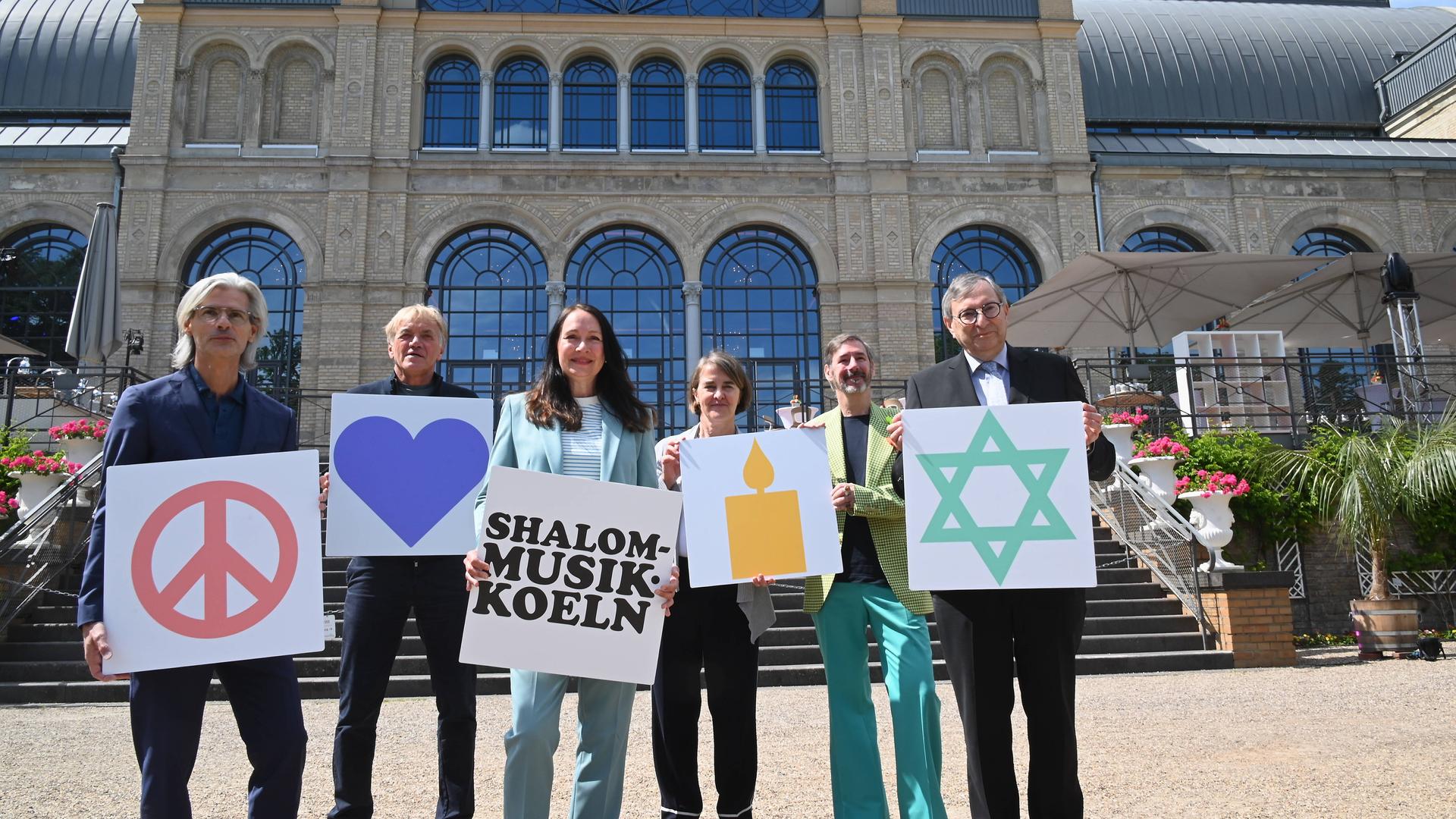 Mehrere Menschen stehen nebeneinander und halten Schilder mit Symbolen und Texten hoch. Auf einem zu lesen steht "Shalom Musik Koeln", auf einem anderen ist ein Davidstern zu sehen.