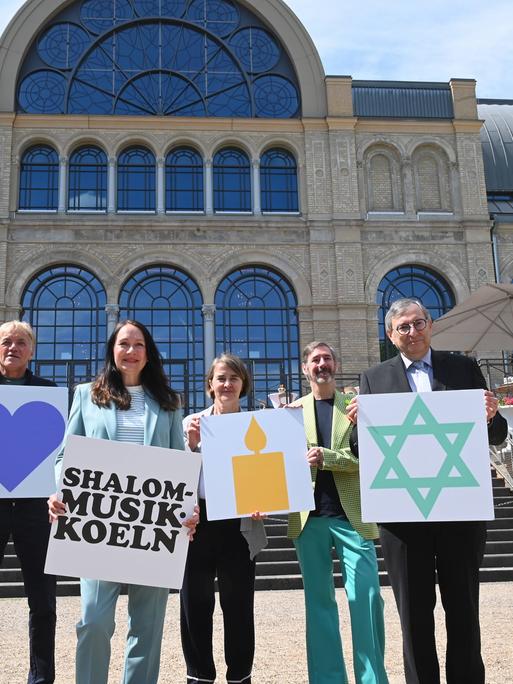 Mehrere Menschen stehen nebeneinander und halten Schilder mit Symbolen und Texten hoch. Auf einem zu lesen steht "Shalom Musik Koeln", auf einem anderen ist ein Davidstern zu sehen.
