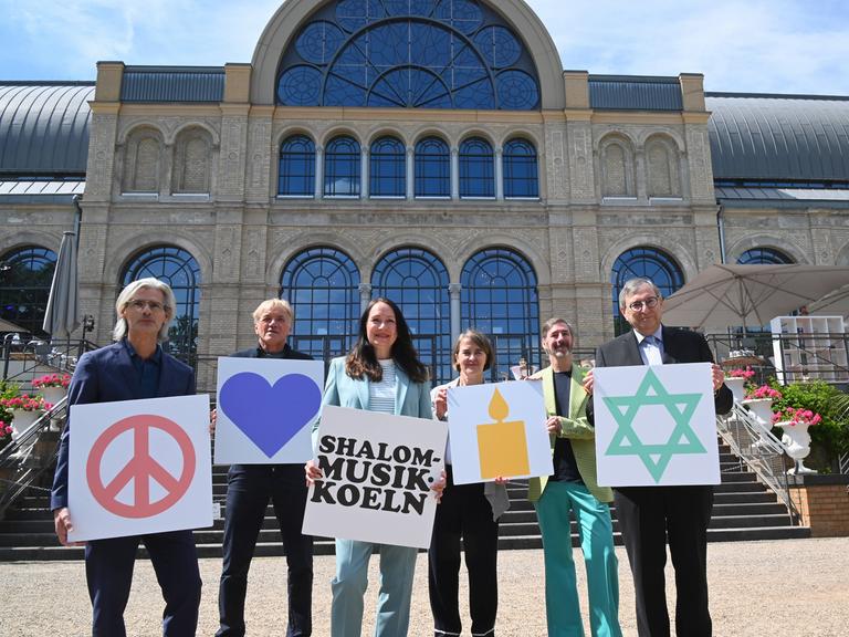 Mehrere Menschen stehen nebeneinander und halten Schilder mit Symbolen und Texten hoch. Auf einem zu lesen steht "Shalom Musik Koeln", auf einem anderen ist ein Davidstern zu sehen.