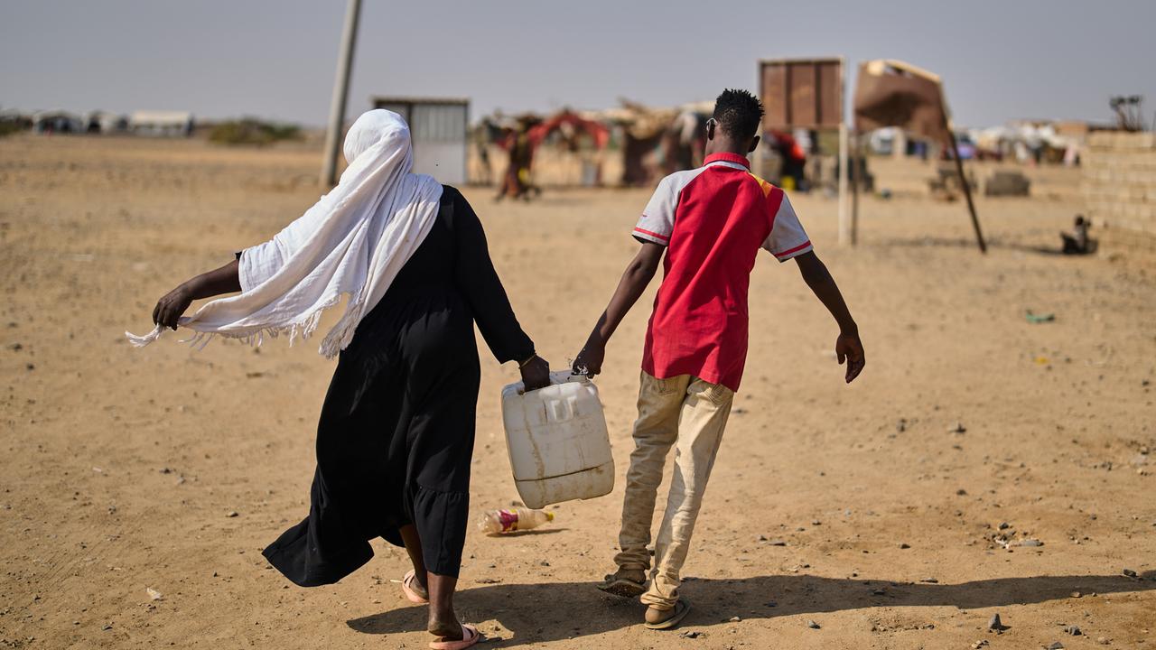 Eine Frau und ein Mann sind in der Rückenansicht in einem Flüchtlingscamp Nahe Port Sudan zu sehen und tragen gemeinsam einen mit Wasser gefüllten Kanister.