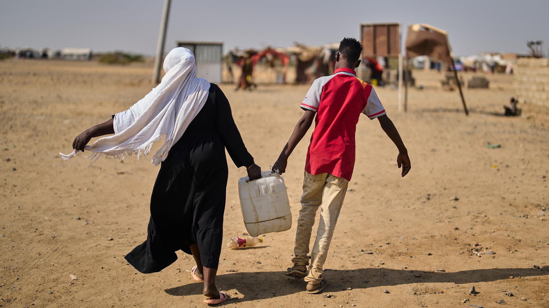 Eine Frau und ein Mann sind in der Rückenansicht in einem Flüchtlingscamp Nahe Port Sudan zu sehen und tragen gemeinsam einen mit Wasser gefüllten Kanister.