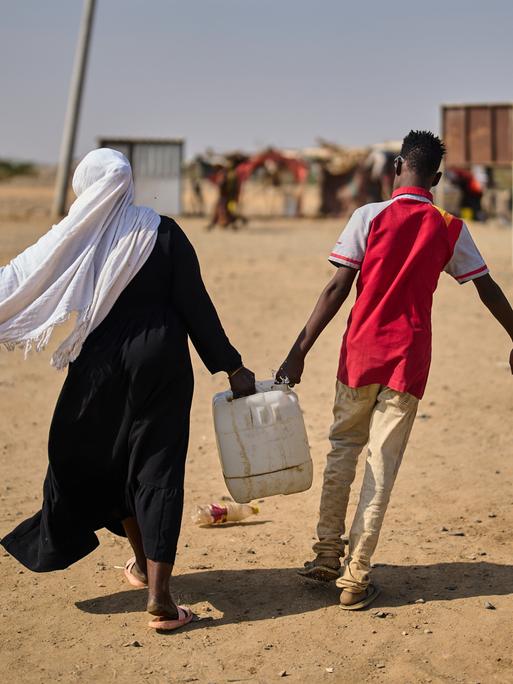 Eine Frau und ein Mann sind in der Rückenansicht in einem Flüchtlingscamp Nahe Port Sudan zu sehen und tragen gemeinsam einen mit Wasser gefüllten Kanister.