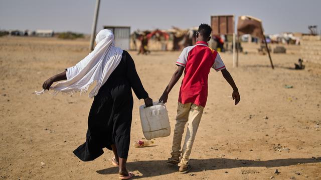 Eine Frau und ein Mann sind in der Rückenansicht in einem Flüchtlingscamp Nahe Port Sudan zu sehen und tragen gemeinsam einen mit Wasser gefüllten Kanister.