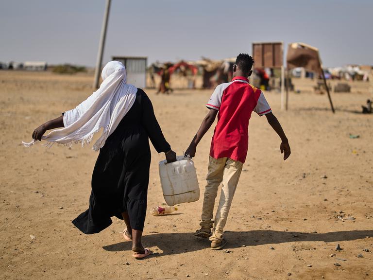 Eine Frau und ein Mann sind in der Rückenansicht in einem Flüchtlingscamp Nahe Port Sudan zu sehen und tragen gemeinsam einen mit Wasser gefüllten Kanister.