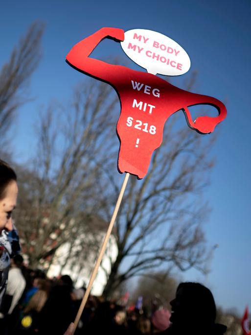 Demonstrantin mit Schild "Weg mit Paragraph § 218 My Body My Choice" und Symbol Uterus 