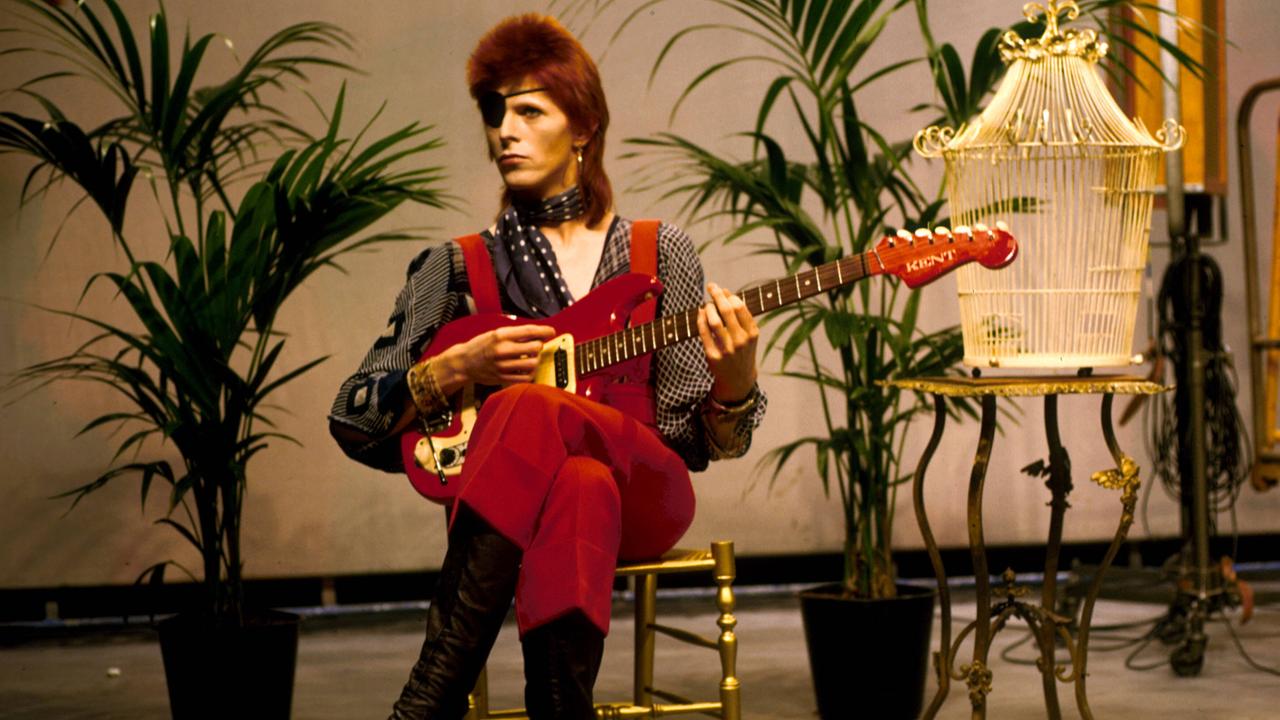 David Bowie in extravaganter Kostümierung. Der Popstar sitzt mit Augenklappe auf einem Stuhl neben einem Vogelkäfig und spielt Gitarre