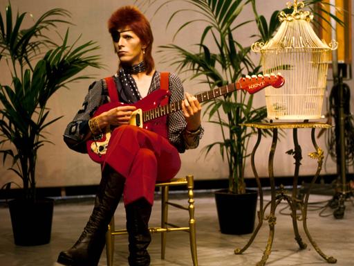 David Bowie in extravaganter Kostümierung. Der Popstar sitzt mit Augenklappe auf einem Stuhl neben einem Vogelkäfig und spiel Gitarre