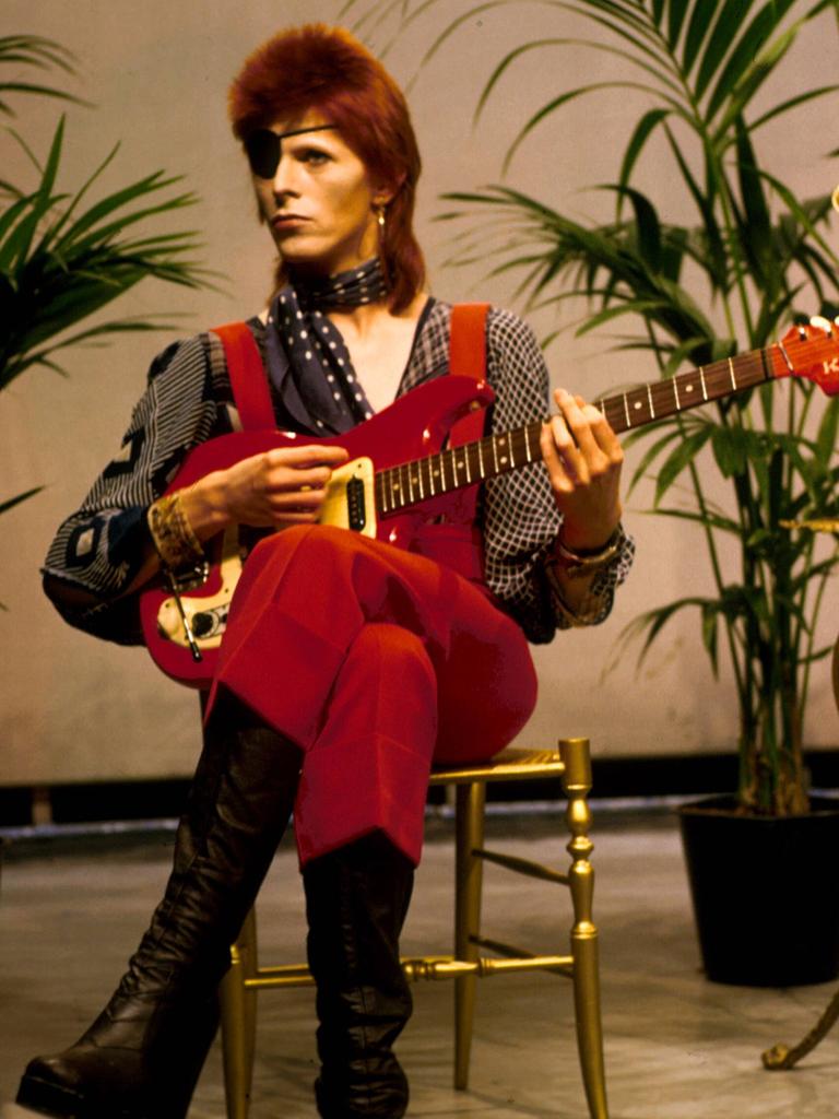 David Bowie in extravaganter Kostümierung. Der Popstar sitzt mit Augenklappe auf einem Stuhl neben einem Vogelkäfig und spiel Gitarre