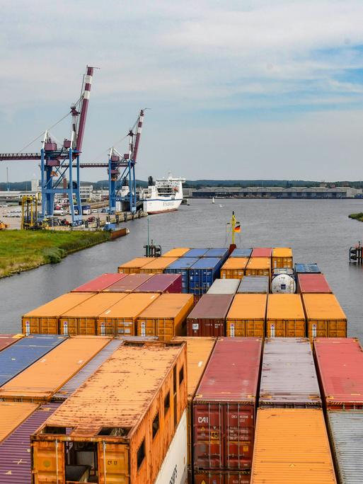 Im Vordergrund sind Container auf einem Frachtschiff zu sehen, im Hintergrund Kräme am Ufer sowie die Trave, auf der das Schiff fährt.