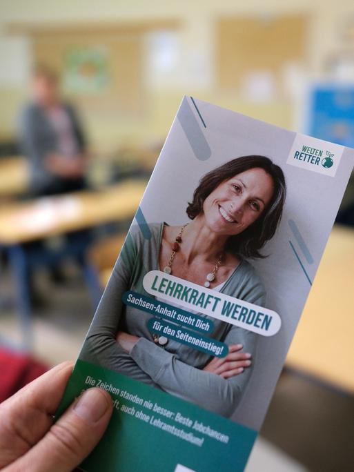 Ein Flyer mit Informationsmaterial zur Berufswahl des Lehrers/der Lehrerin wird in einem Klassenzimmer in die Kamera gehalten. 