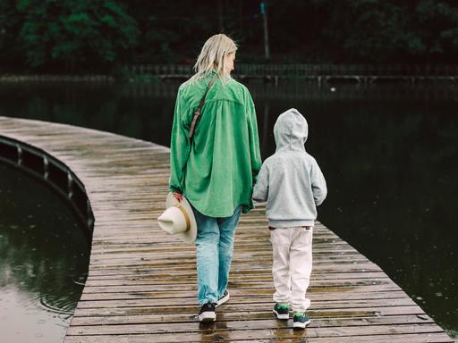 Eine Mutter in grüner Jacke mit ihrem Sohn von hinten auf einem Bootssteg, der über einen See führt. 