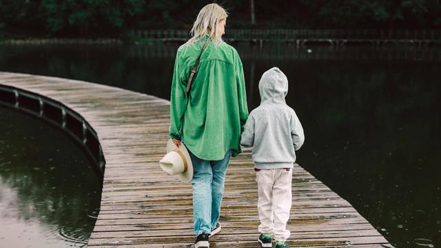 Eine Mutter in grüner Jacke mit ihrem Sohn von hinten auf einem Bootssteg, der über einen See führt. 