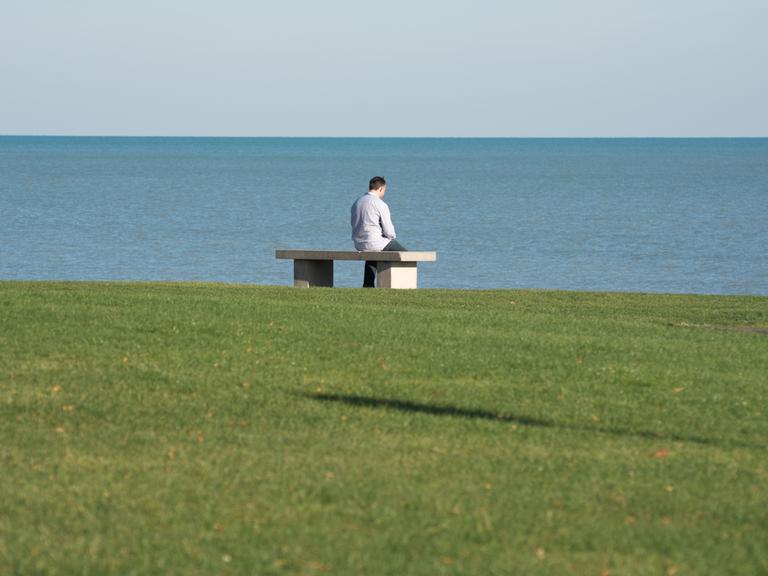 Eine Person sitzt allein auf einer Bank mit leicht gesenktem Kopf vor dem Meer.