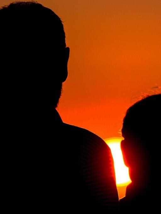 Vater und Sohn stehen mit dem Rücken zur Kamera und betrachten den Sonnenuntergang. 
