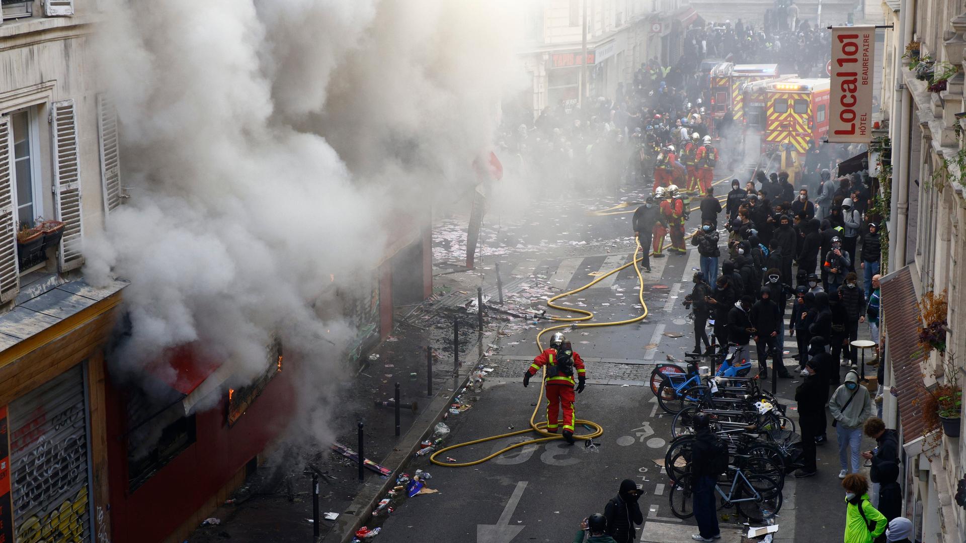 Das Bild zeigt Protestierende und Einsatzkräfte in einer Straße. Aus einem Gebäude quillt dichter Rauch. In der Mitte der Straße sind Feuerwehrleute zu erkennen mit einem langen Schlauch.