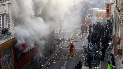 Das Bild zeigt Protestierende und Einsatzkräfte in einer Straße. Aus einem Gebäude quillt dichter Rauch. In der Mitte der Straße sind Feuerwehrleute zu erkennen mit einem langen Schlauch.