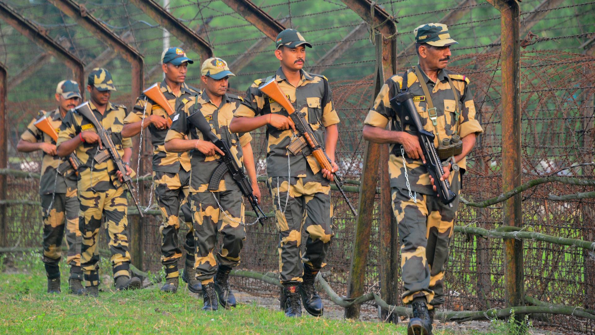 Mehrere bewaffnete indische Soldaten gehen in einer Reihe an einem hohen Grenzzaun entlang. Sie tragen Uniformen, und Mützen. Im Hintergrund sind Bäume zu sehen, die Stimmung wirkt angespannt.