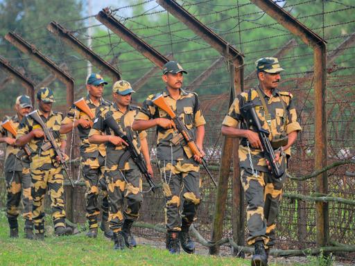 Mehrere bewaffnete indische Soldaten gehen in einer Reihe an einem hohen Grenzzaun entlang. Sie tragen Uniformen, und Mützen. Im Hintergrund sind Bäume zu sehen, die Stimmung wirkt angespannt.