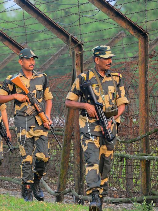Mehrere bewaffnete indische Soldaten gehen in einer Reihe an einem hohen Grenzzaun entlang. Sie tragen Uniformen, und Mützen. Im Hintergrund sind Bäume zu sehen, die Stimmung wirkt angespannt.