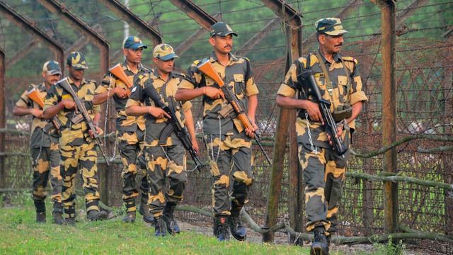 Mehrere bewaffnete indische Soldaten gehen in einer Reihe an einem hohen Grenzzaun entlang. Sie tragen Uniformen, und Mützen. Im Hintergrund sind Bäume zu sehen, die Stimmung wirkt angespannt.