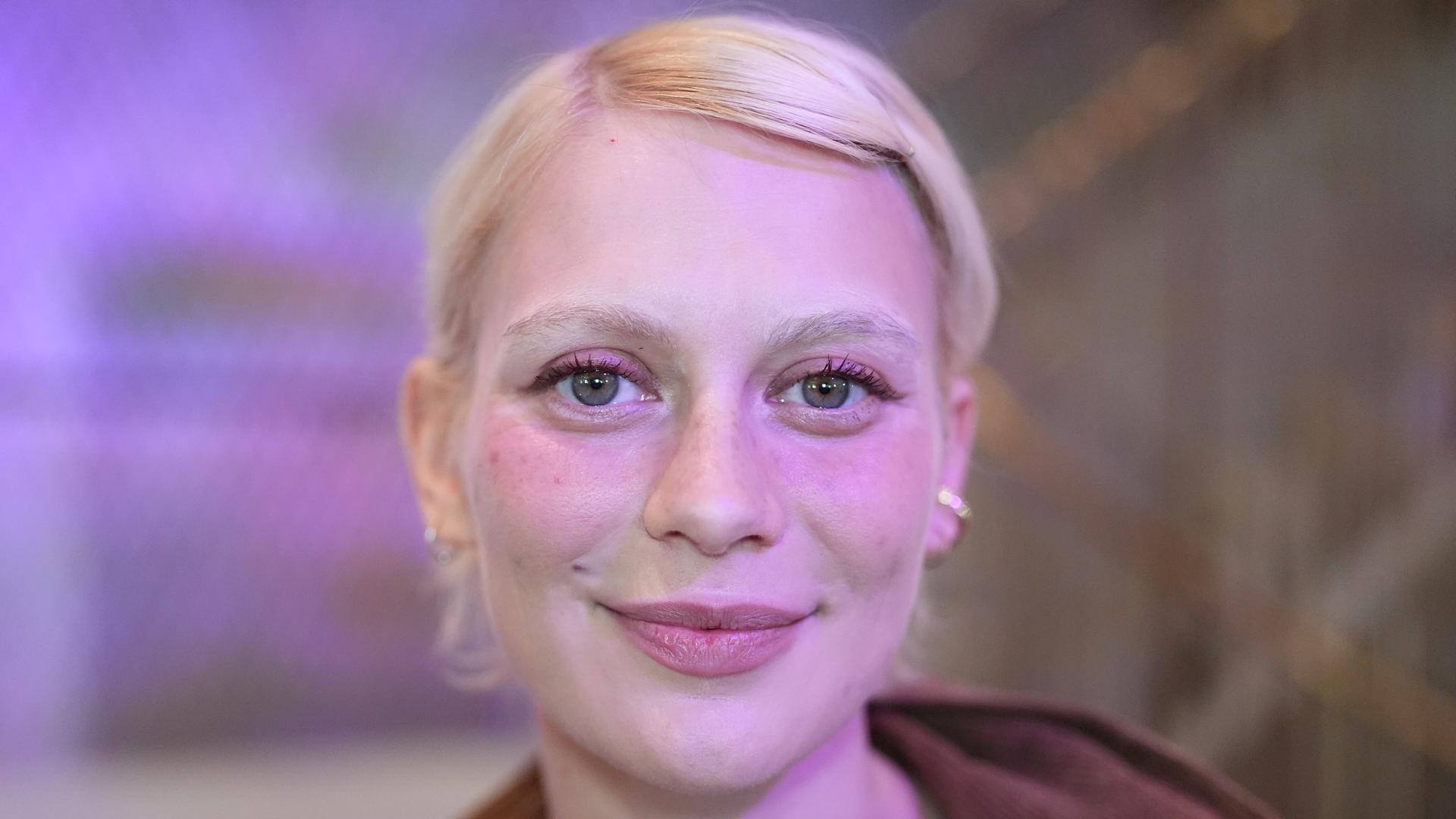 Alli Neumann trägt rosa Lidschatten, steht vor einem violett-rosa beleuchteten Hintergrund und strahlt in die Kamera.