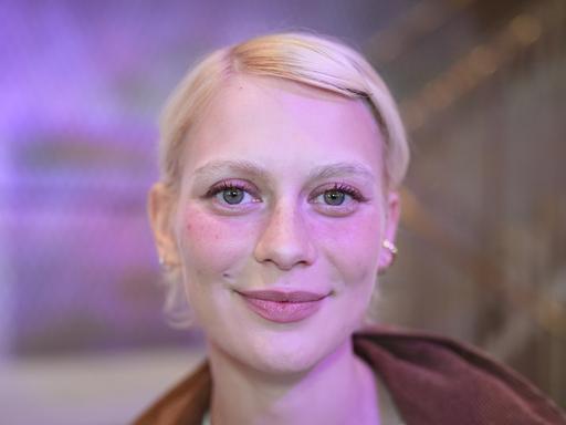 Alli Neumann trägt rosa Lidschatten, steht vor einem violett-rosa beleuchteten Hintergrund und strahlt in die Kamera.