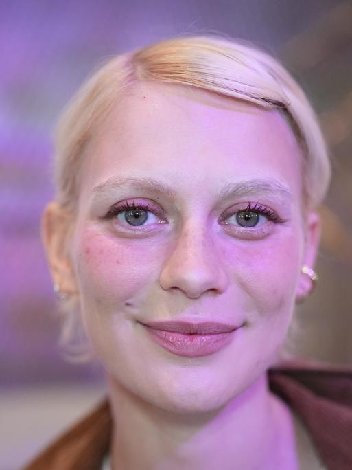 Alli Neumann trägt rosa Lidschatten, steht vor einem violett-rosa beleuchteten Hintergrund und strahlt in die Kamera.