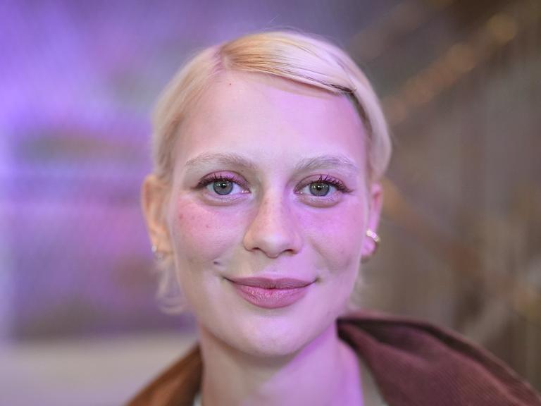 Alli Neumann trägt rosa Lidschatten, steht vor einem violett-rosa beleuchteten Hintergrund und strahlt in die Kamera.