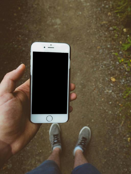 Eine Person blickt von oben auf die Füße, sie hält ein Smartphone in der Hand, dessen Bildschirm schwarz ist. Eine Person blickt von oben auf die Füße, sie hält ein Smartphone in der Hand, dessen Bildschirm schwarz ist.