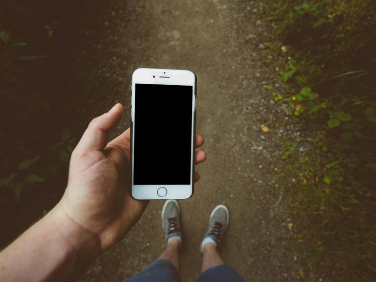 Eine Person blickt von oben auf die Füße, sie hält ein Smartphone in der Hand, dessen Bildschirm schwarz ist. Eine Person blickt von oben auf die Füße, sie hält ein Smartphone in der Hand, dessen Bildschirm schwarz ist.