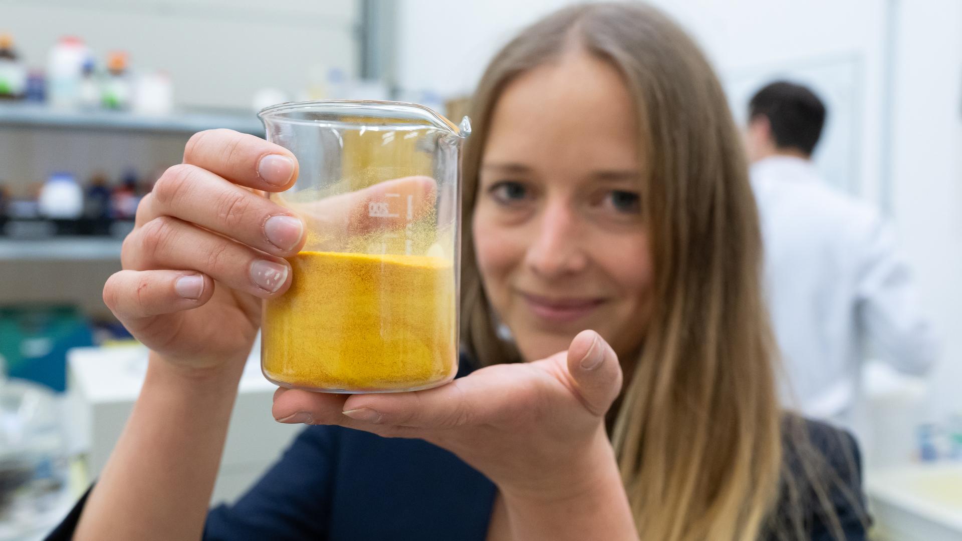 Anne Lamp steht in einem Labor und hält ein Gefäß mit einer gelben, schlierigen Substanz in die Kamera.