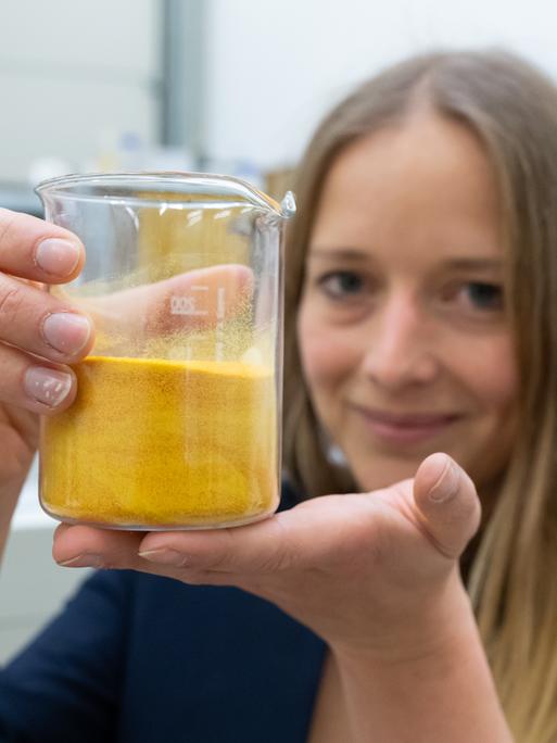 Anne Lamp steht in einem Labor und hält ein Gefäß mit einer gelben, schlierigen Substanz in die Kamera.
