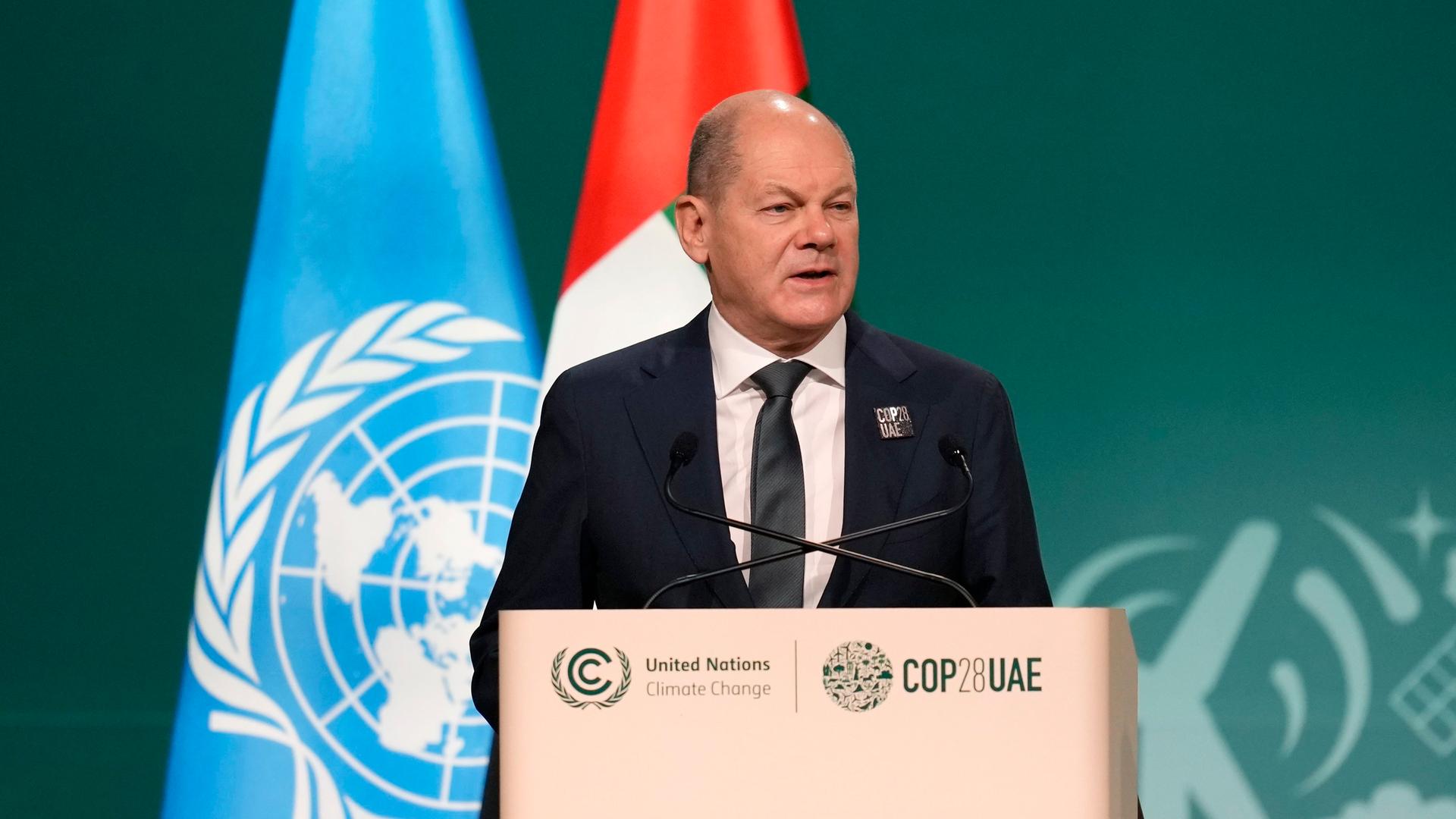 Bundeskanzler Scholz steht bei der COP28 am Rednerpult, hinter ihm die Flagge der UNO und der Vereinigten Arabischen Emirate.