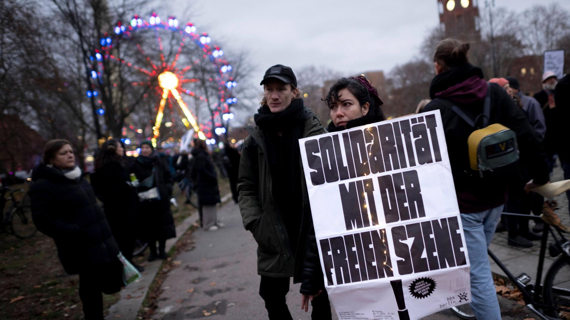 Ein Mann und eine Frau stehen am Rand einer Demonstration. In der Hand hält die Frau ein Schild. Die Aufschrift: "Solidarität mit der Freien Szene."