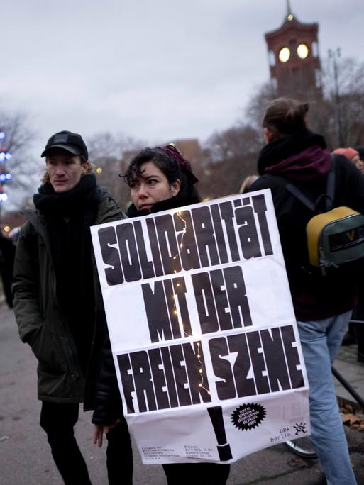 Ein Mann und eine Frau stehen am Rand einer Demonstration. In der Hand hält die Frau ein Schild. Die Aufschrift: "Solidarität mit der Freien Szene."