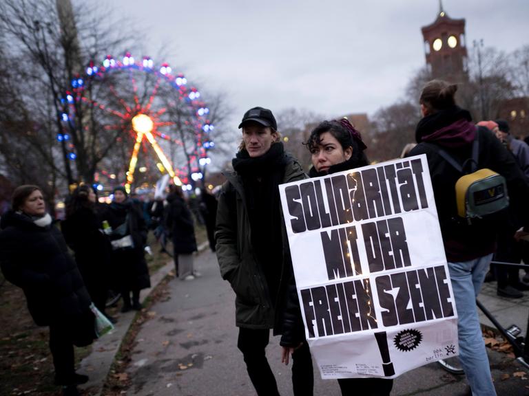 Ein Mann und eine Frau stehen am Rand einer Demonstration. In der Hand hält die Frau ein Schild. Die Aufschrift: "Solidarität mit der Freien Szene."